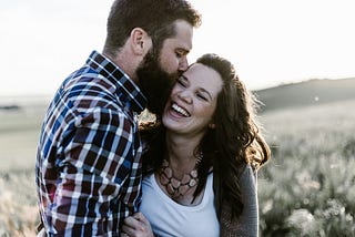 A man kisses a laughing woman in a crop field, both radiating happiness amidst nature.