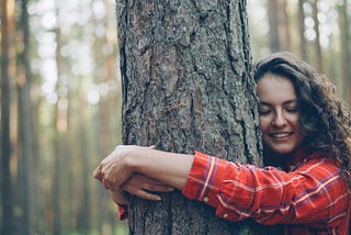 a woman in a red plaid shirt encircles a tree trunk iwth her arms and presses her face against the bark