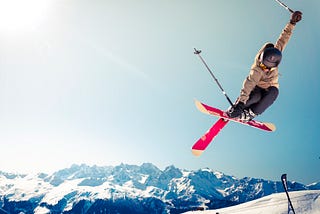 airborne skiier crosses red skis midair with snowy countains in background