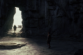 A distant figure strides out of an ancient rock cave towards a crack that leads out towards the sun.
