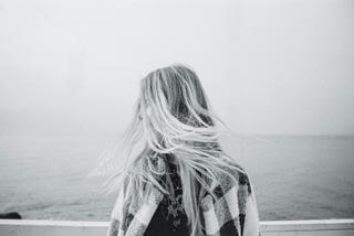 A black and white image of a woman facing the ocean with wind in her hair