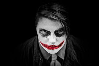 A person with face paint resembling a dark, intense clown or joker character, with white makeup covering the face, dark black circles around the eyes, and a wide red, smudged smile painted across the mouth. The individual stares directly into the camera with a piercing gaze, creating a haunting and dramatic effect against a black background.