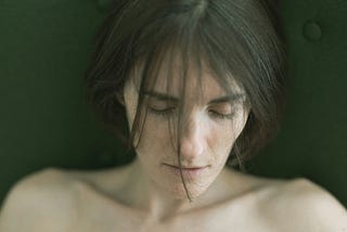 a naked woman with closed eyes, her soft features framed by tousled hair against a muted green backdrop. She is probably dead