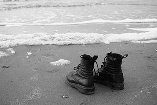 A black-and-white photograph of an empty pair of black shoe-boots at the edge of calm ocean water at the shore