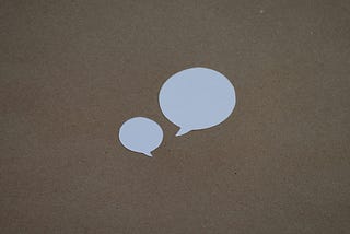 Two voice bubbles cut out of white paper on a brown background. One is small and the other large to illustrate the idea of the copywriting technique of starting short.
