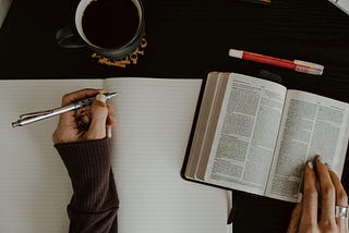 A left-handed person holding a pen over a notebook with a book beside it