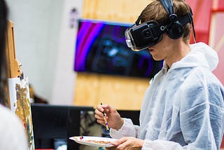 A person wearing a VR headset holds a paintbrush and palette in front of a canvas.