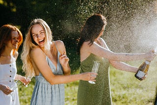 3 pretty women are having fun splashing liquid from a bottle in a greenery area. KoolShooters/Pexels.