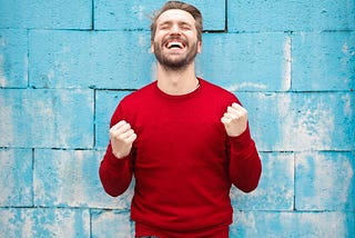 A happy guy standing by the wall in red shirt with his eyes closed, fists rising in air, and a broad smile.