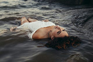 A woman in a white dress floating peacefully in a body of water with her eyes closed and a serene expression, suggesting a moment of reflection or release. Her dark, curly hair fans out around her, and the water around her ripples gently, evoking a sense of calm and perhaps symbolizing a poignant moment of letting go or healing.