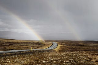 Photo of a road winding through open moorland with a grey sky and two rainbows in the distance.