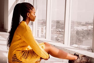 A brown girl sits sadly on a tiled white floor in front of a tall white outlined window. As she wears a yellow dress.