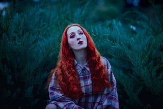 A white woman with David Bowie-esque bright pink and red makeup and long orange curly hair, wearing a black, pink, and white flannel and standing before a fern/bush.