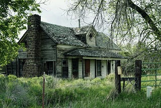 An abandoned house with dark windows and overgrown grass rising up the walls.