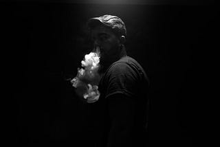 A man in the dark vaping a large puff of vapor