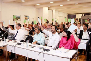 A group of mostly white people raise white slips of paper. In the front row, two brown people are not raising papers, and appear unhappy.