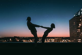 Silhouette of a couple on a nighttime rooftop, holding hands and leaning back from each other; she looks as though she may fall if he lets go.