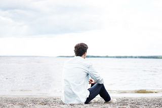 Man sitting on the beach looking at the water, casually dressed in white shirt, jeans, and tennis shoes.