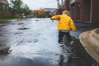 A small child wearing a yellow coat jumping into a puddle on an empty street.