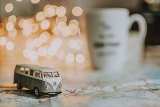 a toy VW campervan on a map on a table, in the background, out of focus, are fairy lights and a mug.