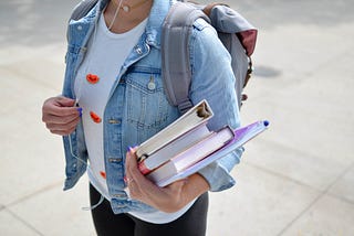 Girl, wearing jean jacket carries books in left arm, while wearing a backpack and holding headphone wires in her right hand.