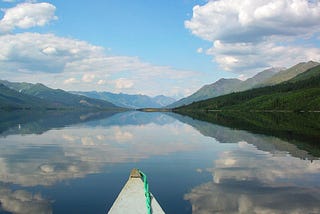 View of a lake from a boat or canoe. There are mountains in the background and a partly cloudy sky above.