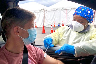 A white man wearing a salmon colored shirt in a car and wearing a blue surgical mask is receiving a vaccine through the window from a Black nurse wearing scrubs, blue gloves, an N95 masks, and a blue handkerchief with polka dots.
