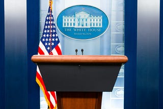 Image of a podium next to a US flag with the White House emblem behind it.