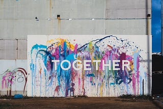 A large colourful canvas stands against a white wall, with the word TOGETHER superimposed over it in large white capital letters.
