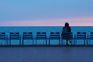 A woman sitting alone among empty chairs facing the ocean at sunset, looking out quietly