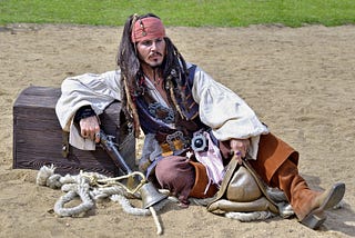 A man is dressed as famous pirate character ‘Captian Jack Sparrow’. He is lying down on sand, leaning against a wooden chest, holding an old pistol.