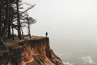 A person stands at the edge of a forest near a cliff’s edge overlooking a fog covered sea.