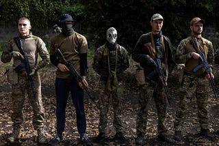 Five Ukrainian men stand lined up at attention with weapons in their hands dressed in mixed camo.