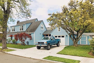 A two-story home painted blue with an attached garage. A blue pickup truck is parked in the drive. The grass and landscaping are very tidy.