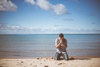 A man kneeling in prayer in front of a body of water.