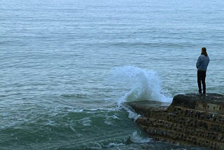 A man standing at the end of a pier with waves crashing against the rock wall