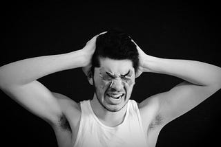 A man in a white cotton vest has his hands on his head, eyes covered and appears to be screaming