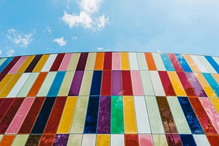 Photo of a wall with vertical rectangles placed next to each other of different colors, forming a colorful wall. Above it, you can see a blue sky with some clouds.