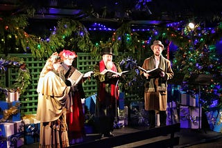 Three Carolers with song books in hand and decrative lights in the background.