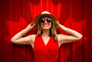A woman smiling wearing a red dress standing in front of a red wall. Her hands are holding a hat on her head and she is wearing sunglasses and gold jewelry.