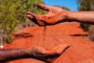 One Aborignal person's hand pours red soil from into another Aboriginal person'’ hand in an arid landscape with sparse vegetation and a clear blue sky, symbolizing a connection to the land.