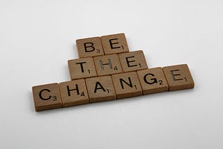 Scrabble tiles spelling out “be the change” on a white background.