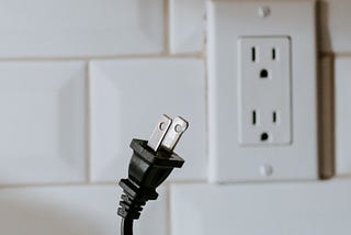 Black electrical plug pulled from a white wall socket on a white tiled wall.
