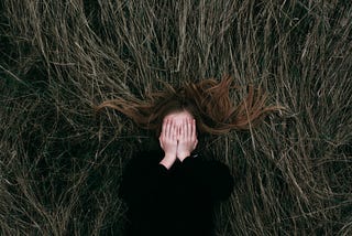 Woman lies in a field, covering her faces with her hands.