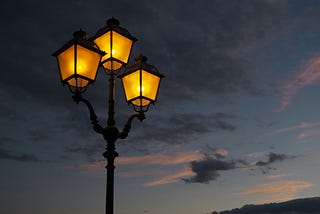 A lamp post with three lanterns, each glowing yellow, against a lightly clouded sky at twilight
