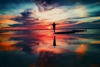 Person standing with outstretched arms looking at a colorful sky over the ocean