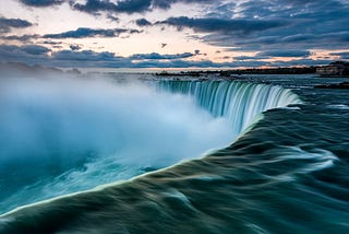 Niagara Falls from a side view. Deep blue water and cloudy skies.