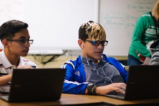 This image shows two students sitting in a classroom with laptops in front of them.