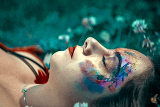 A freelancer lies on grass with eyes closed, colorful paint adorning their face. White flowers are scattered around, and the background is blurred. The scene exudes a serene, artistic atmosphere, capturing the essence of a solopreneur’s tranquil escape from failing systems.