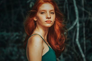 red haired woman in a green spaghetti strapped dress, staring at the camera. Behind here are pale branches against a dark background